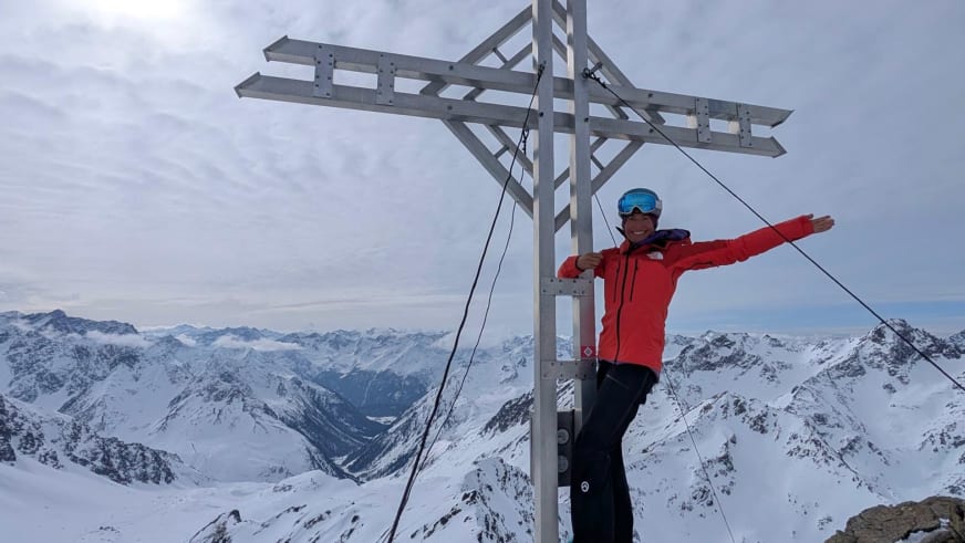 Bergführerin Monika Hümmer in roter Winterjacke mit ausgebreiteten Armen vor Holzgipfelkreuz.
