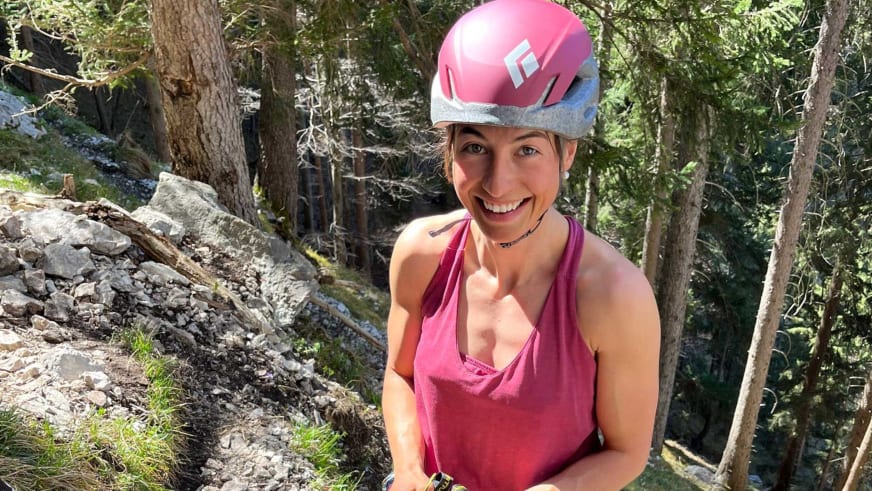 Bergführerin Monika Hümmer mit rosa Kletterhelm und Klettergurt zeigt den Daumen hoch vor Felsen und Waldvegetation.