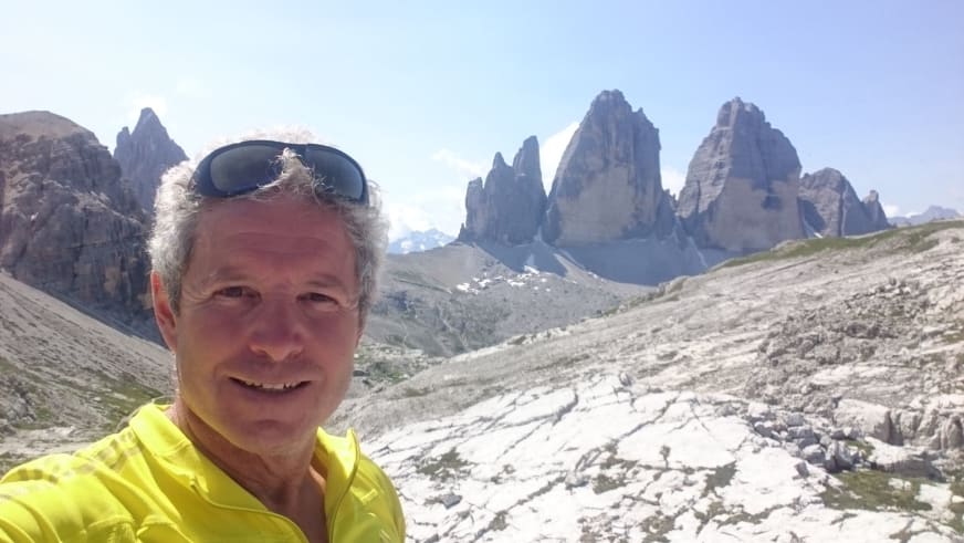 Bergführer Jürgen Bäumler macht Selfie in gelber Jacke vor hellen Kalksteintürmen der Dolomiten.