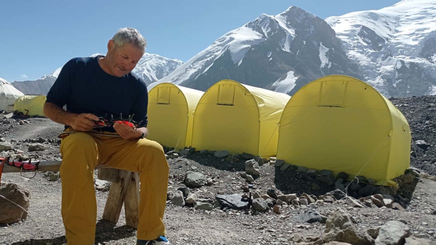 Bergführer Jürgen Bäumler sitzt auf Holzblock im Hochgebirgslager zwischen gelben Expeditionszelten.