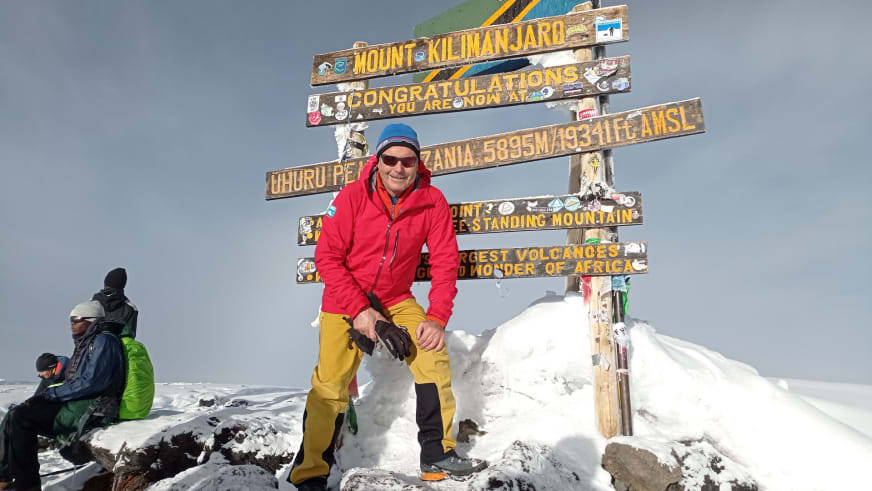 Bergführer Jürgen Bäumler steht in roter Daunenjacke am Uhuru Peak Schild des Mount Kilimanjaro auf 5895 Metern.