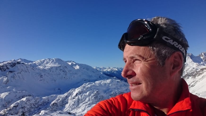 In roter Softshell-Jacke und mit Skibrille posiert Bergführer Jürgen Bäumler vor verschneiten Gipfeln.