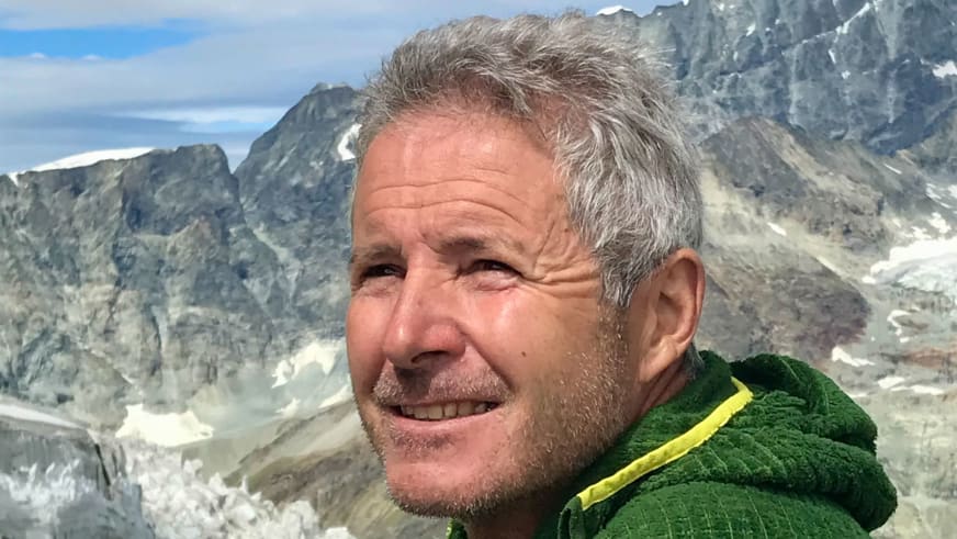 Bergführer Jürgen Bäumler lächelt in grüner Softshelljacke vor hochalpiner Berglandschaft mit Schneefeldern.
