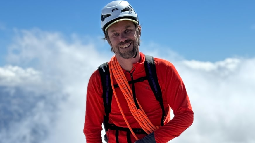 Bergführer Christoph Vonmetz lächelt in die Kamera und trägt weißen Kletterhelm, rote Jacke und oranges Seil.