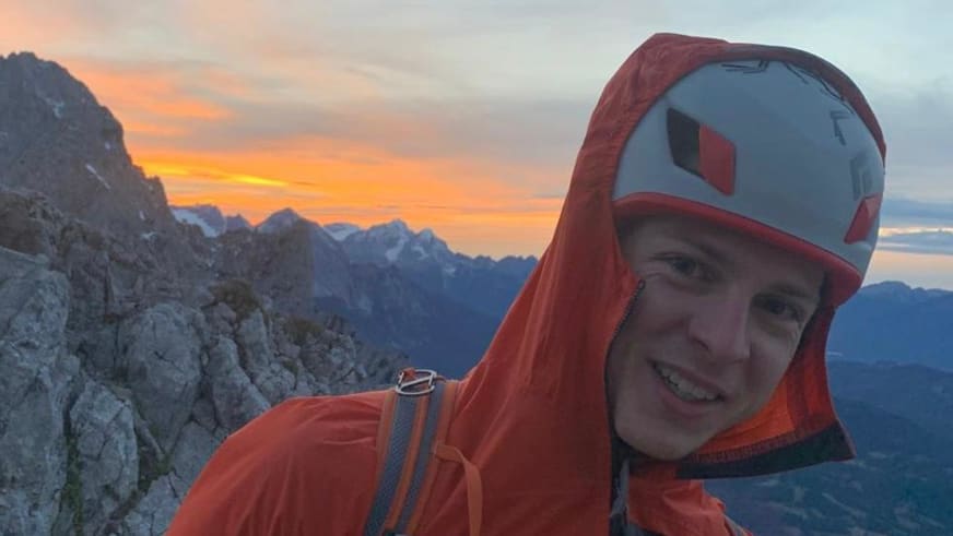 Bergführer Wolfgang Wagner lächelt in die Kamera, trägt orange Softshelljacke und Helm vor Bergkulisse im Morgenlicht.