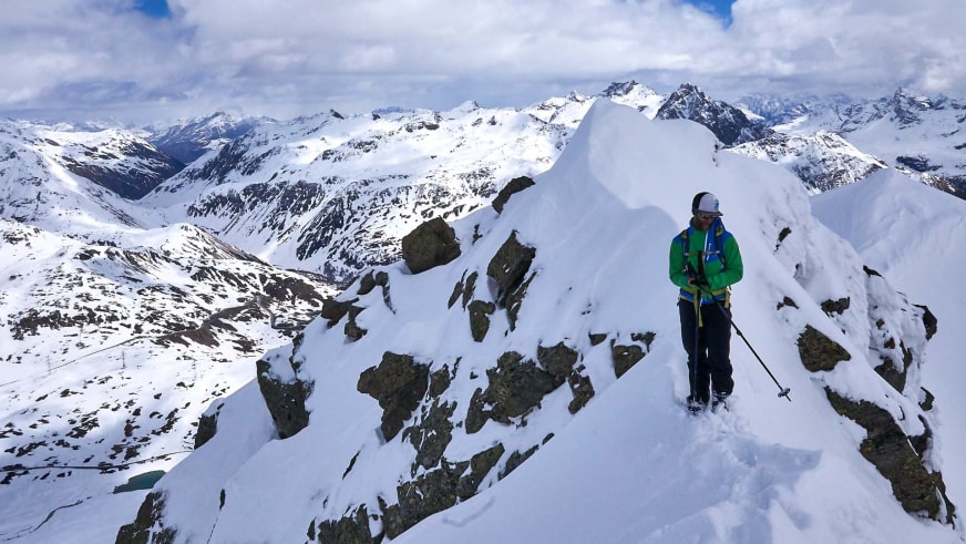 Bergführer Simon Taffner steht in grüner Winterjacke und weißem Helm auf einem schmalen, verschneiten Felsgrat im Hochgebirge.