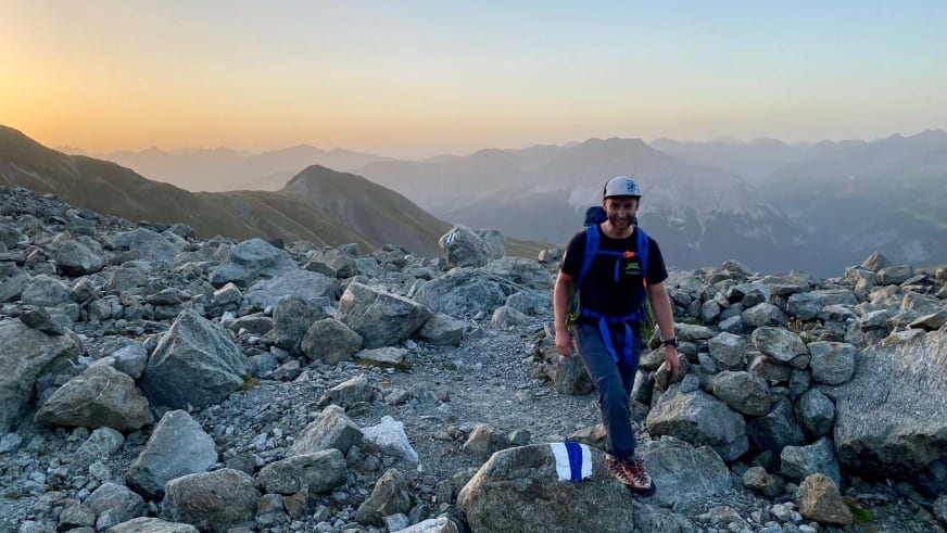 Auf einem Geröllfeld aus grauen Felsblöcken steht Bergführer Simon Taffner in dunkler Kleidung mit weißer Kappe und blauem Rucksack.