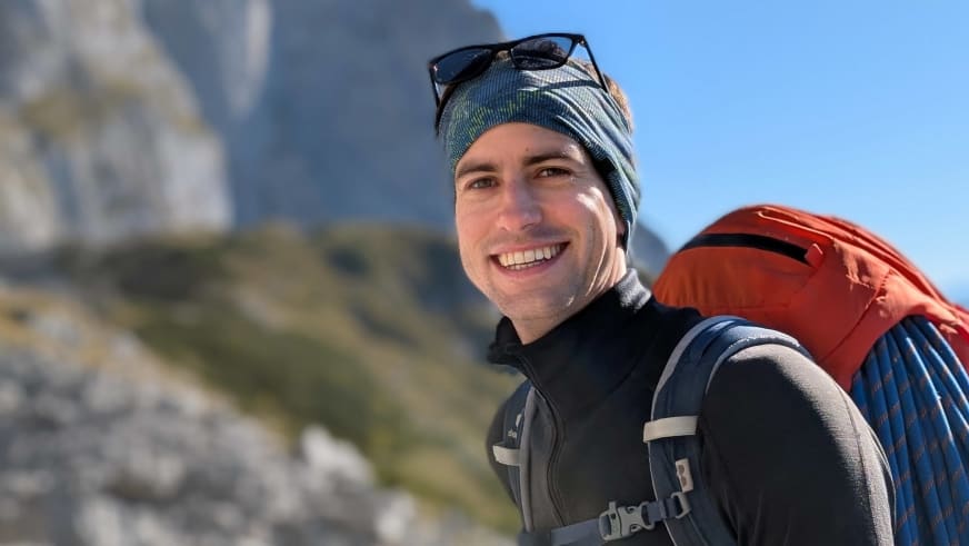 Lächelnder Bergführer Christoph Meysel mit gestreifter Mütze und orangem Rucksack vor Felswänden.