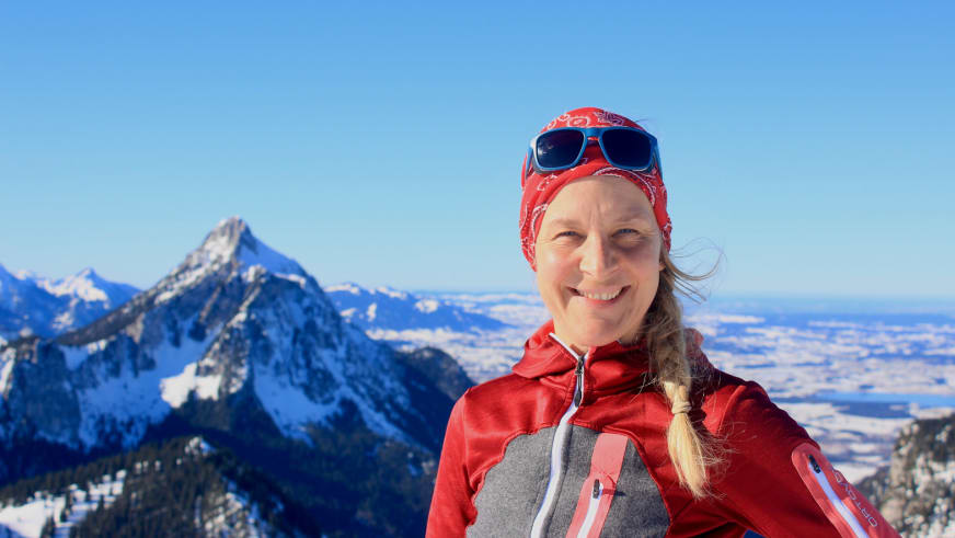 Portrait zeigt Bergwanderführerin Bernadette Schnabl lächelnd in roter Softshelljacke mit rotem Bandana vor verschneiten Bergen.
