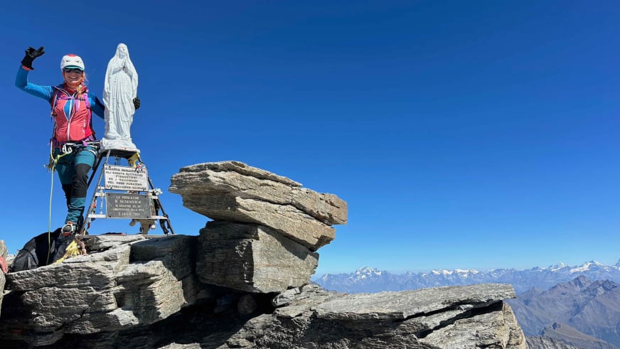 Bergwanderführerin Bernadette Schnabl winkt am Gipfel mit weißer Marienstatue, trägt rote Jacke und weißen Helm vor Bergpanorama.