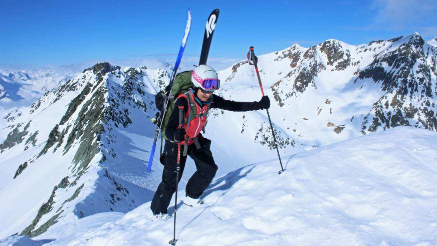 Bergwanderführerin Bernadette Schnabl steigt auf Skiern durch Pulverschnee auf, trägt weißen Helm und rot-schwarze Jacke vor Schneegipfeln.