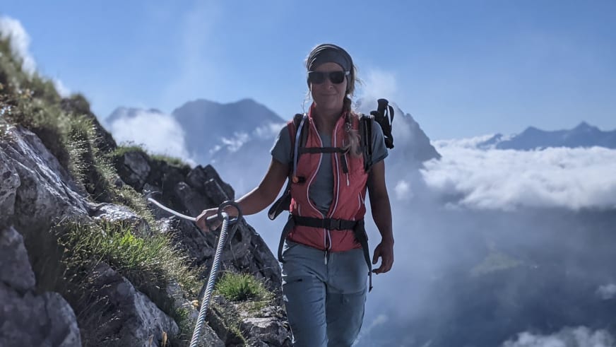 Bergwanderführerin Bernadette Schnabl hält sich am Stahlseil fest und geht schmalen Bergpfad mit roter Weste und blauer Hose.