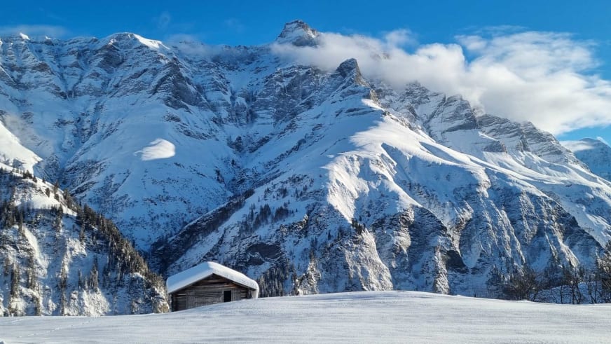 Traditionelle Holzhütte in verschneiter Landschaft des Safientals nahe Hotel Camana mit Bergpanorama der Adula-Alpen.