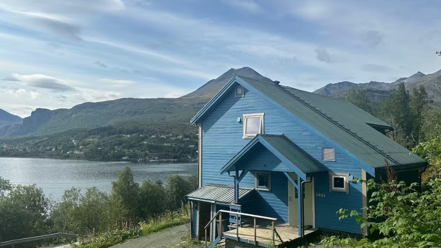 Das Lyngen Blue House liegt erhöht über einem Fjord mit bewaldeten Bergen im Hintergrund.