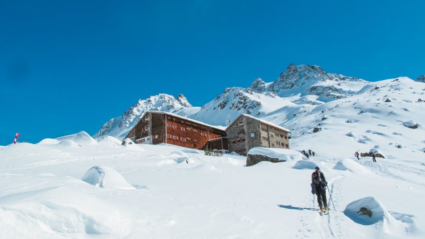 Skitourengeher vor der verschneiten Jamtalhütte in der Silvretta bei klarem Wetter.
