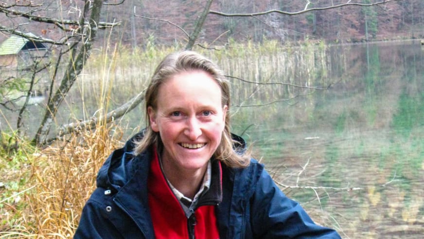 Portrait zeigt Bergwanderführerin Christine Brandmüller mit blonden Haaren und dunkelblauer Outdoor-Jacke vor einem herbstlichen See.