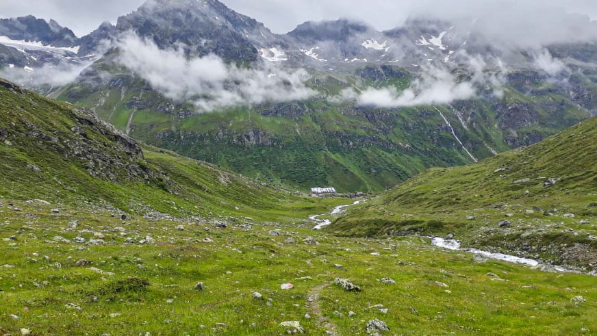 Sommeraufnahme der Jamtalhütte im grünen Jamtal mit schneebedeckten Berggipfeln im Hintergrund.