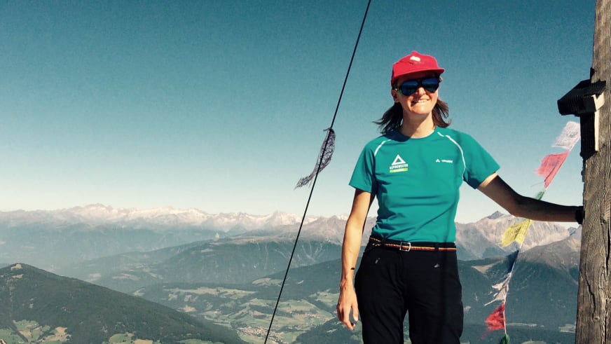 Bergwanderführerin Christine Brandmüller steht lächelnd am hölzernen Gipfelkreuz mit bunten Gebetsfahnen in den Dolomiten.