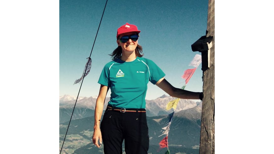 Auf der Zugspitze trägt Bergwanderführerin Christine Brandmüller rote Kappe und türkises T-Shirt neben dem Holzkreuz mit Gebetsfahnen.