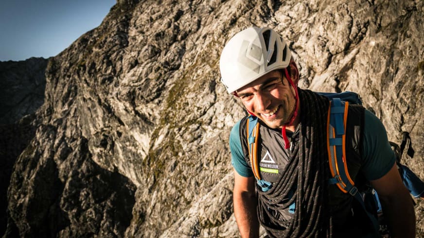Bergführer Josef Neumeier mit weißem Helm und blauer Kleidung lächelt vor einer steilen Kalksteinfelswand.