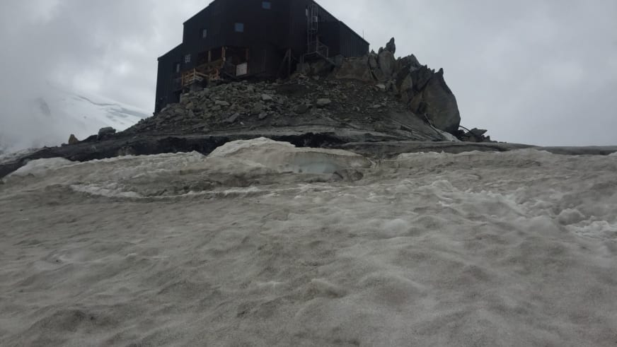 Das Refuge des Cosmiques steht auf einem Felsgrat zwischen Firnfeldern bei bewölktem Himmel.