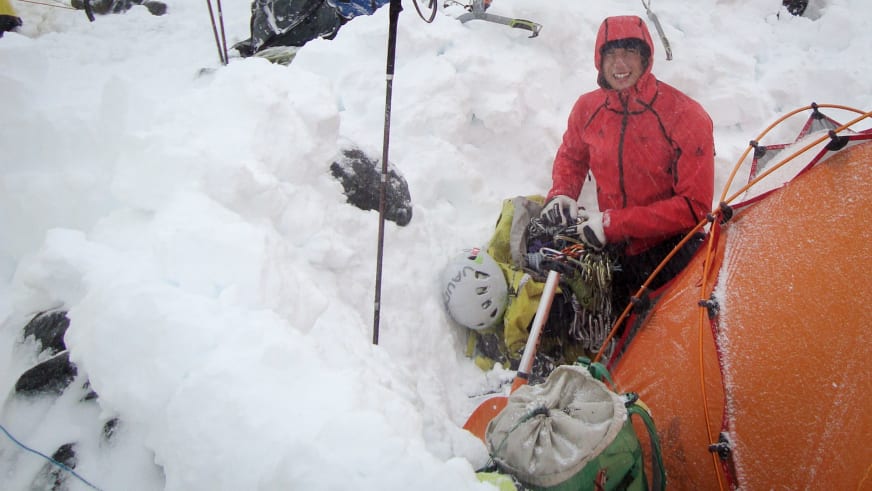 Bei einer Expedition sitzt Bergführer Korbinian Schmidtner in roter Daunenjacke vor dem orangem Zelt im Schnee.