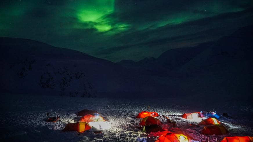 Bergwanderführer Jonas Haass steht bei beleuchteten Expeditionszelten auf schneebedecktem Terrain unter grünen Nordlicht-Schleiern am Nachthimmel.