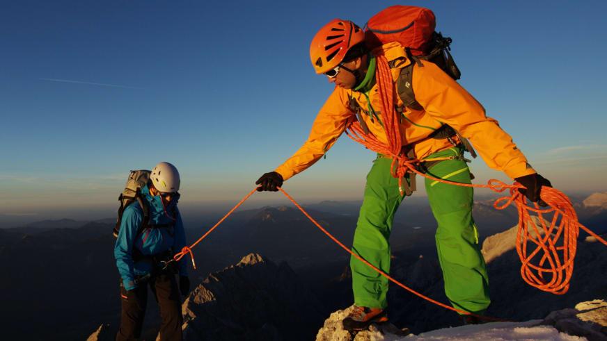 Auf einem schneebedeckten Felsgrat steht Peter Fresia Bergführer mit orangefarbenem Seil und einem weiteren Bergsteiger bei goldenem Abendlicht.