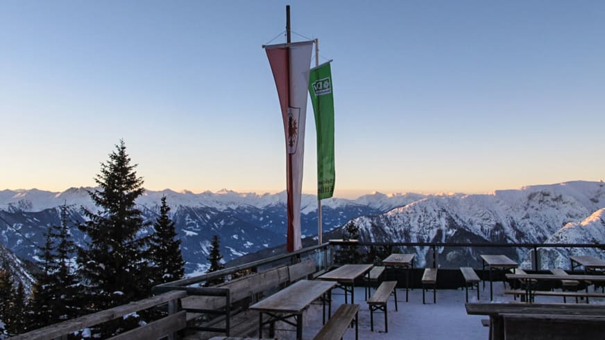 Verschneite Terrasse der Erfurter Hütte mit Holztischen und österreichischen Fahnen vor Alpenpanorama.