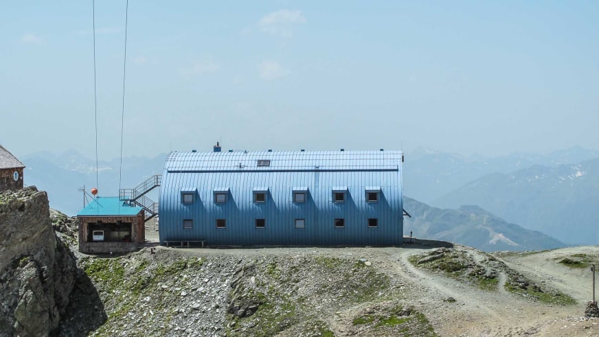 Außenansicht der Stüdlhütte mit blau-grauer Wellblechfassade und symmetrisch angeordneten Fenstern.