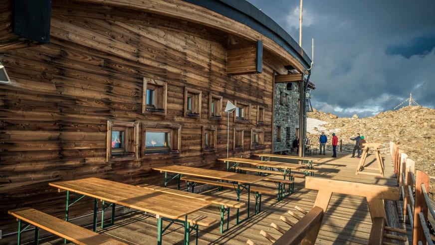 Holzterrasse mit Bierbänken am Rifugio Città di Mantova und Bergsteigern in alpiner Landschaft.