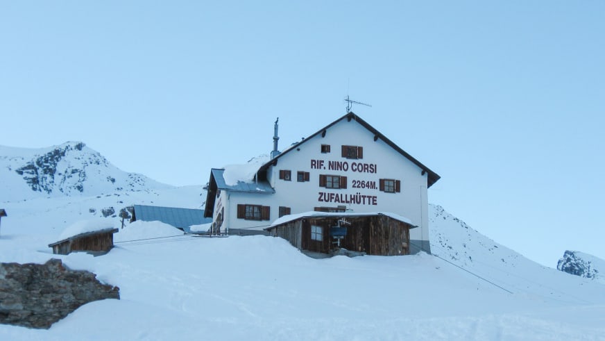 Verschneite Zufallhütte mit weißem Hauptgebäude und Holzanbau in winterlicher Hochgebirgslandschaft.