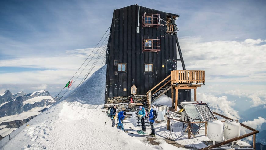 Bergsteiger in bunter Winterkleidung stehen vor der Capanna Margherita auf schneebedecktem Gipfel der Walliser Alpen.