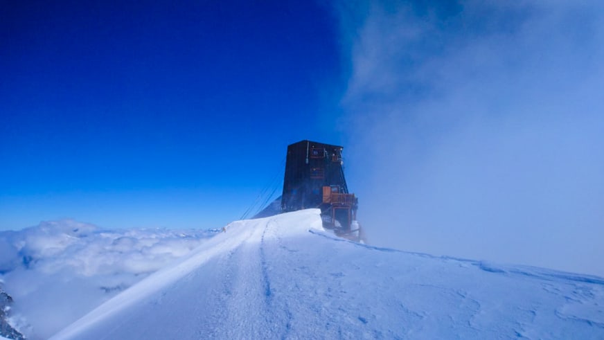 Winteraufnahme zeigt die Capanna Margherita als dunkles Gebäude auf verschneitem Bergrücken.