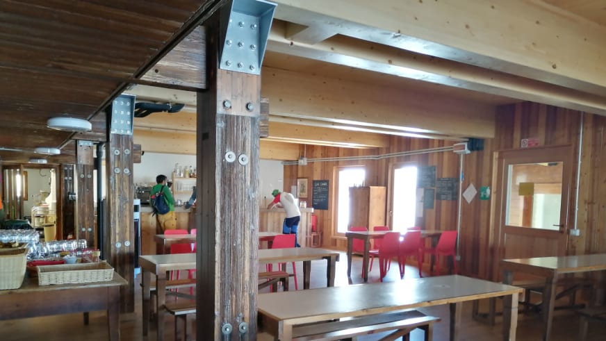 Gastraum des Rifugio Quintino Sella al Felik mit Holztischen und offener Küche in moderner Stahlkonstruktion.