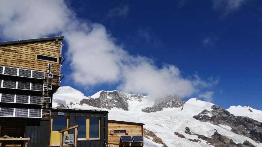 Sicht auf das Rifugio Quintino Sella al Felik mit vergletscherter Bergkette und Solarpaneelen an der Fassade.
