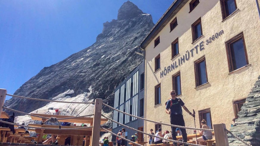 Die Hörnlihütte bei klarem Himmel mit Wanderern auf der Terrasse und steilem Felsgipfel dahinter.