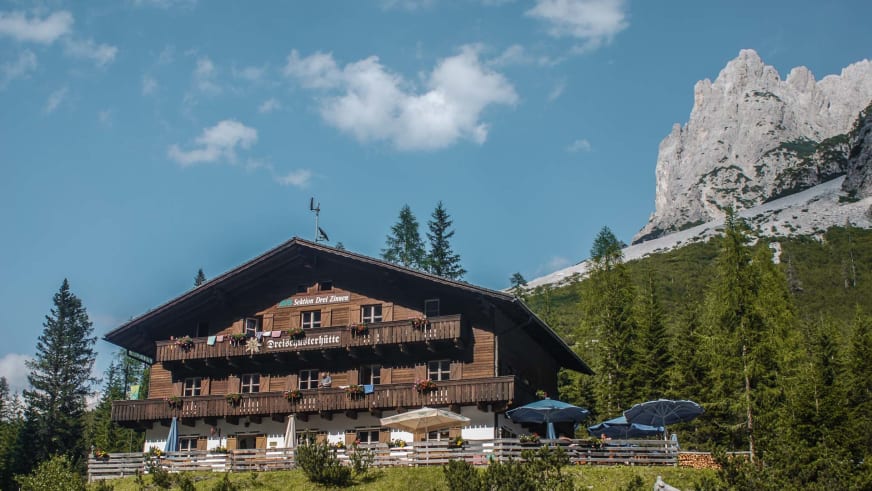 Blick auf die Drei Schuster Hütte mit Holzfassade, Balkonen und Sonnenterrasse vor steilen Felswänden.