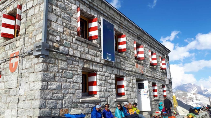 Bergwanderer pausieren vor der Rothornhütte mit rot-weißen Fensterläden vor verschneiter Bergkulisse.