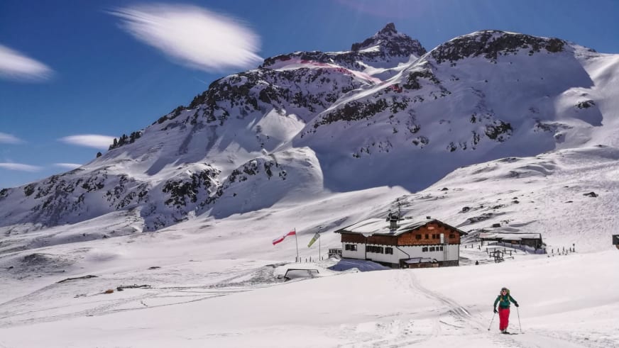 Skifahrer in grüner Jacke fährt durch verschneite Landschaft vor der Sesvenna Hütte mit Bergpanorama.