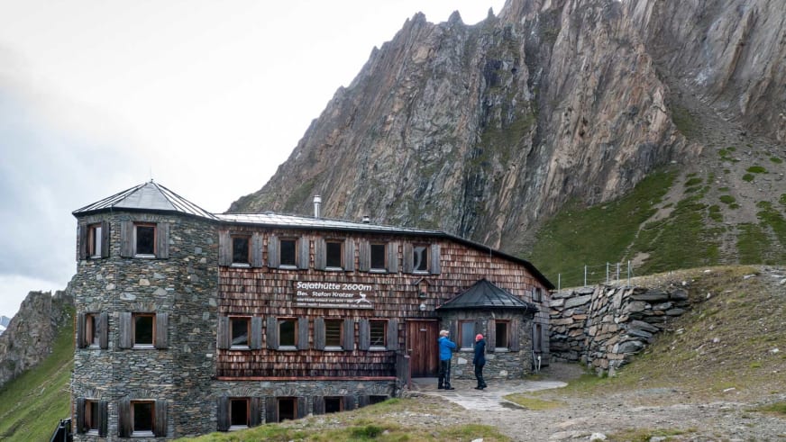 Zwei Wanderer stehen vor der Sajathütte, einer Berghütte aus Naturstein und Holz auf 2.600 m Höhe.