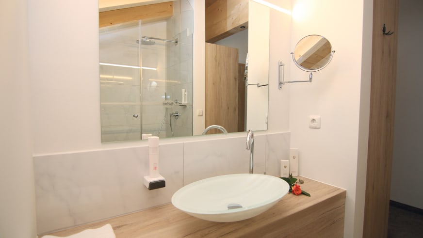 Modernes Hotelbadezimmer im Alpinhotel Berchtesgaden mit weißem Waschbecken und Holzbalken an der Decke.
