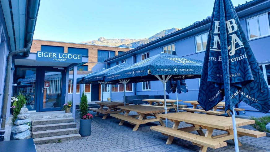 Außengastronomie der Eiger Lodge mit vier Picknicktischen unter blauen Sonnenschirmen vor der Rezeption.