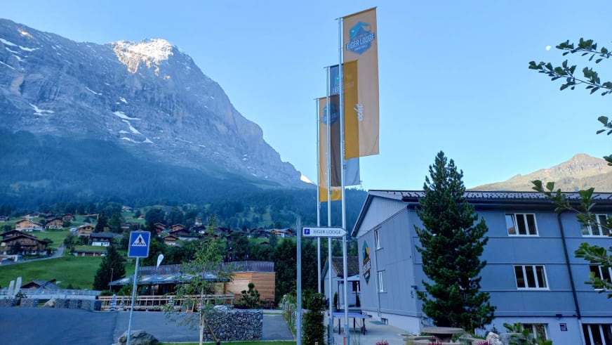 Die Eiger Lodge zeigt sich zur blauen Stunde mit drei Fahnenmasten und schneebedeckten Gipfeln im Hintergrund.