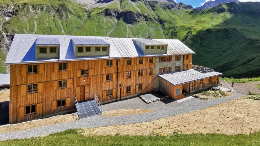 Luftbild der Kemptner Hütte mit mehreren Holzgebäuden und Solarpaneelen auf grüner Almwiese.