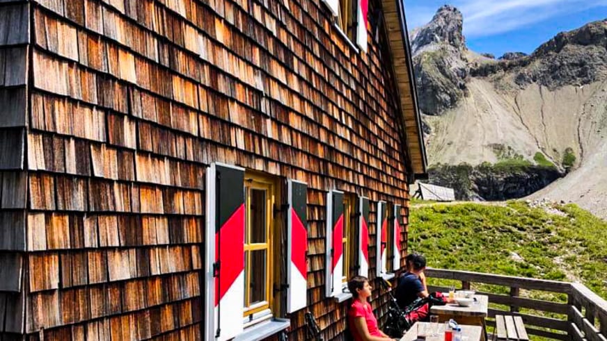 Bergwanderer sitzen auf der Terrasse der Memminger Hütte vor alpinem Bergpanorama bei sonnigem Wetter.