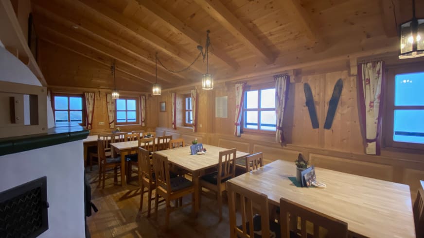Gastraum der Warnsdorfer Hütte mit Holztischen, Kachelofen und rot-weißen Vorhängen.