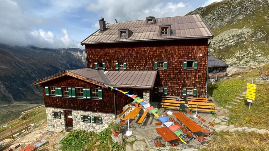 Sonnenterrasse der Warnsdorfer Hütte mit orangefarbenen Holzstühlen vor alpiner Bergkulisse.