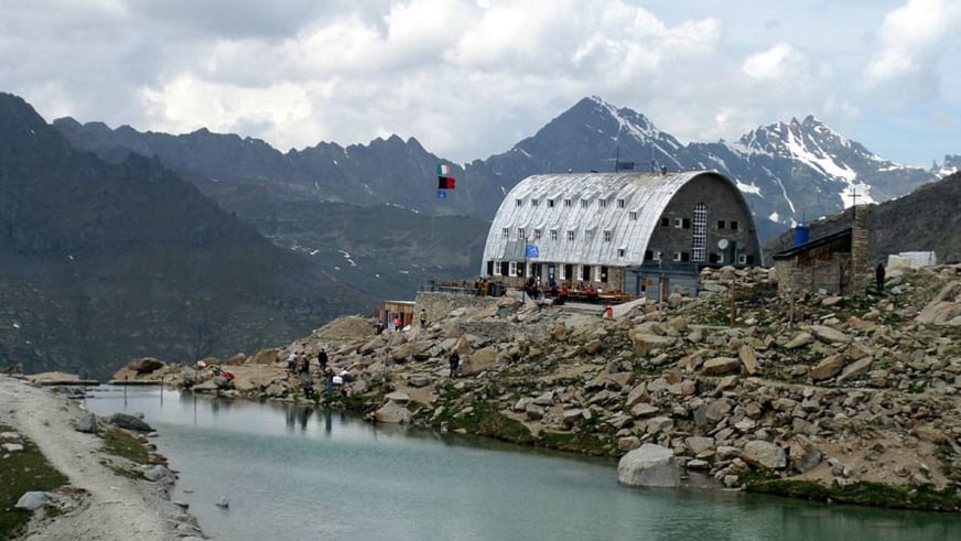 Wanderer vor dem Rifugio Vittorio Emanuele II mit charakteristischem Tonnendach und kleinem Bergsee im Vordergrund.