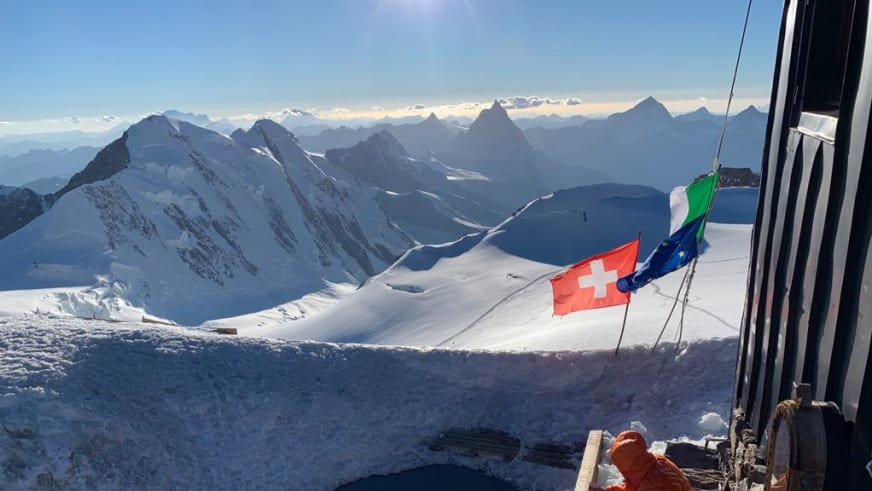 Blick von der Capanna Gnifetti über verschneite Gipfel der Walliser Alpen mit italienischer Fahne.